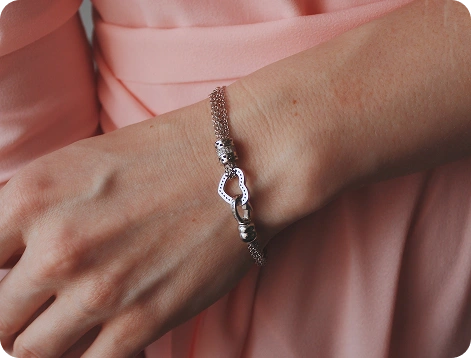 Close-up van een elegante zilveren armband met hartvormige details, gedragen om de pols van een vrouw tegen een zachte roze stof achtergrond.