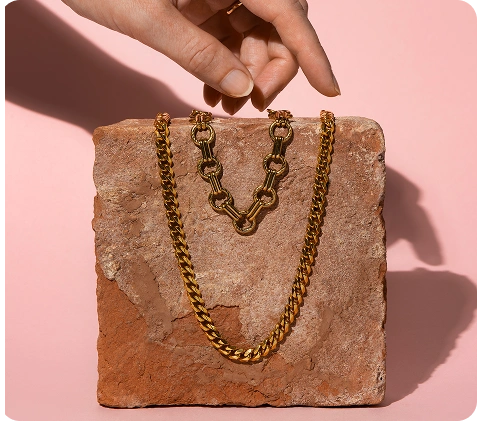 Stijlvolle gouden kettinghalskettingen tentoongesteld op een gestructureerd stenen oppervlak met een hand die naar de juwelen reikt in zacht studiolicht.