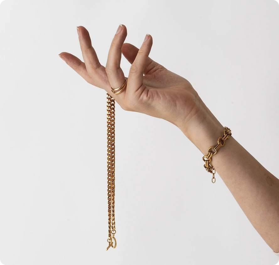 Close-up van de hand van een vrouw die een delicate gouden kettinghalsketting vasthoudt terwijl ze een elegante gouden armband en minimalistische ringen draagt.
