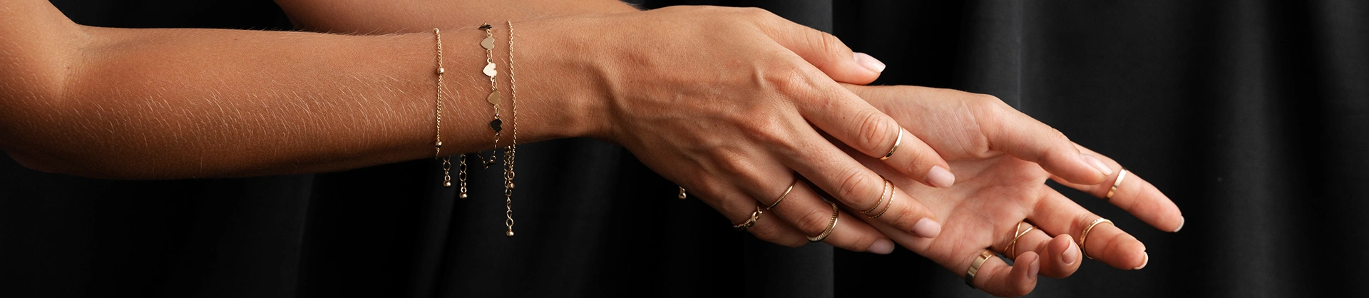 Artistieke close-up van vrouwelijke handen die gelaagde gouden armbanden en minimalistische ringen dragen tegen een donkere elegante achtergrond.